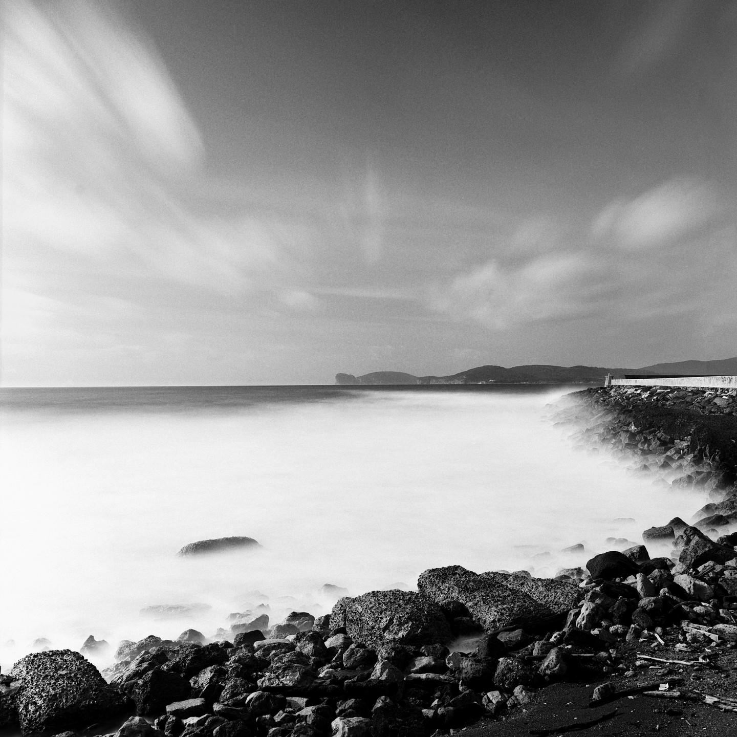 A long exposure captures the movement of water and clouds along a rocky coastline with distant hills.
