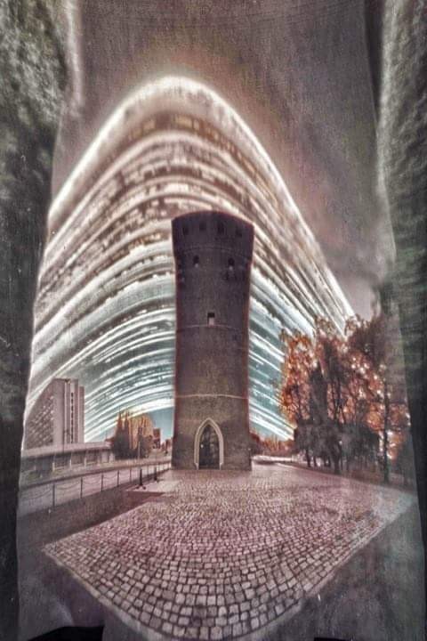 A medieval tower stands on a cobblestone square with a modern building and trees in the background, all under a sky filled with star trails.