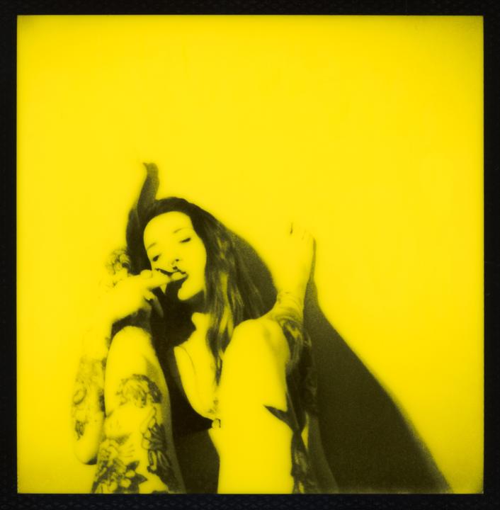 A woman with long hair and tattoos is shown in a yellow and black image, with her fingers near her mouth.