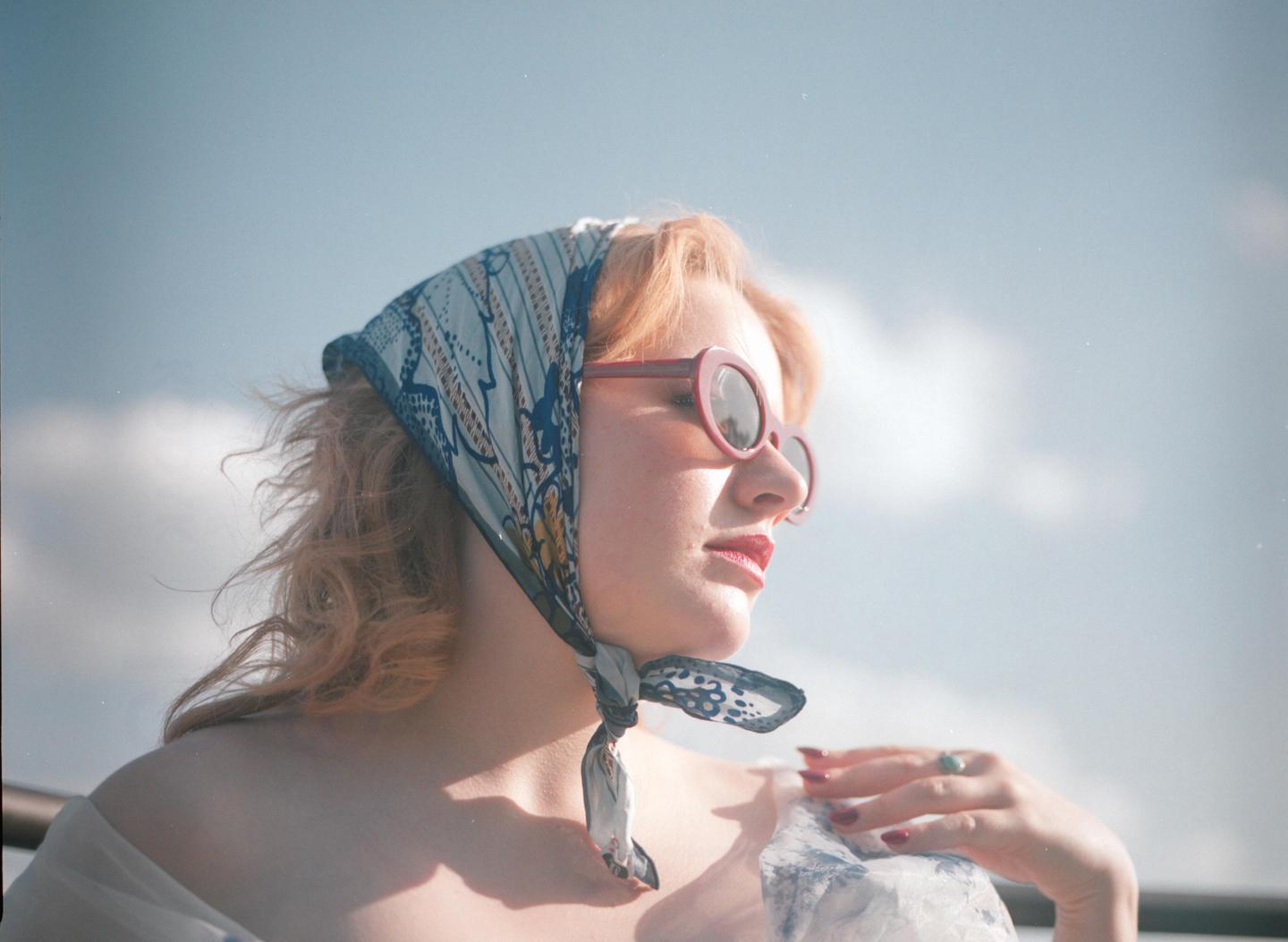 A woman with a headscarf and sunglasses looks to the side against a bright sky.