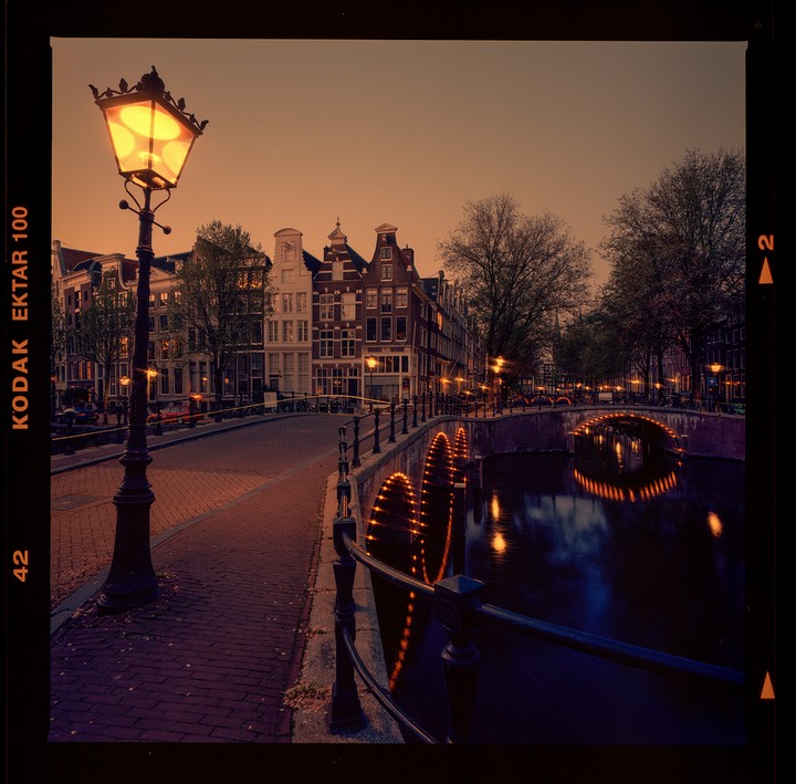 A street lamp illuminates a brick road and canal with bridges and buildings in the background at dusk.