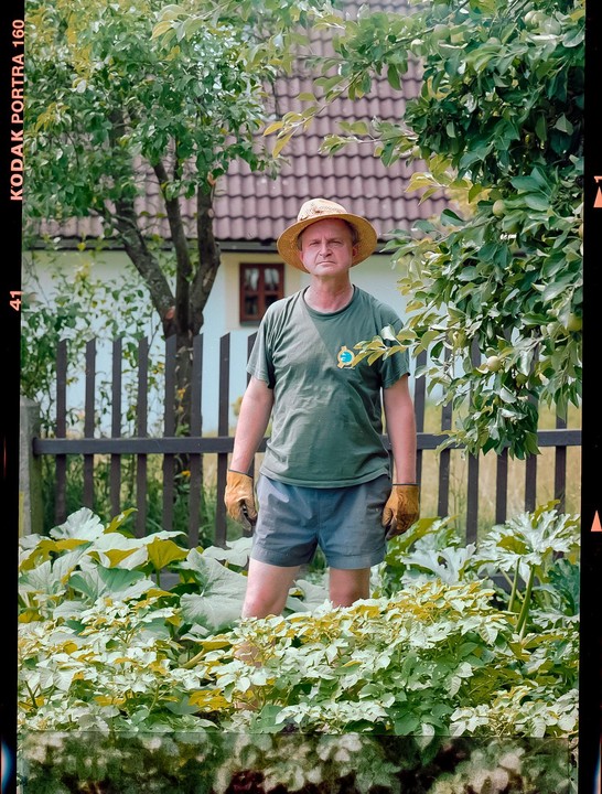 A man wearing a straw hat and gardening gloves stands in a garden with lush plants and a wooden fence behind him.