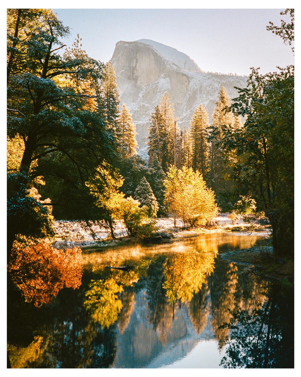 A snow-dusted mountain and forest reflect in a river during golden hour.