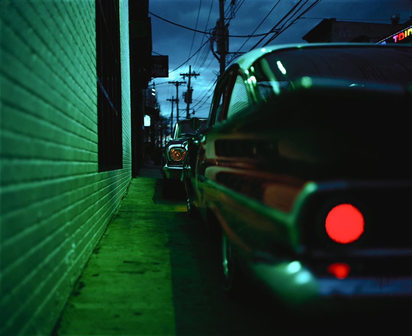 Two vintage cars are parked along a green brick wall at night, with power lines and a neon sign visible in the background.