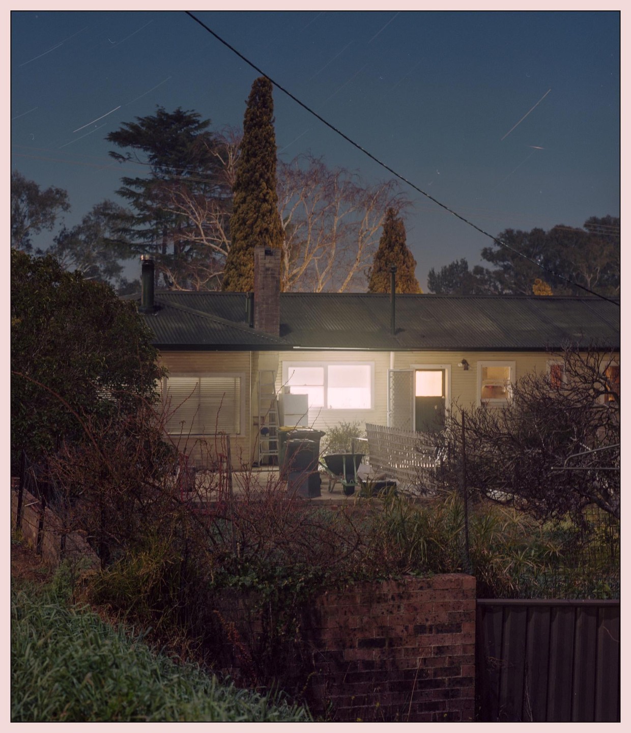 A house with illuminated windows is seen at night under a sky with star trails.
