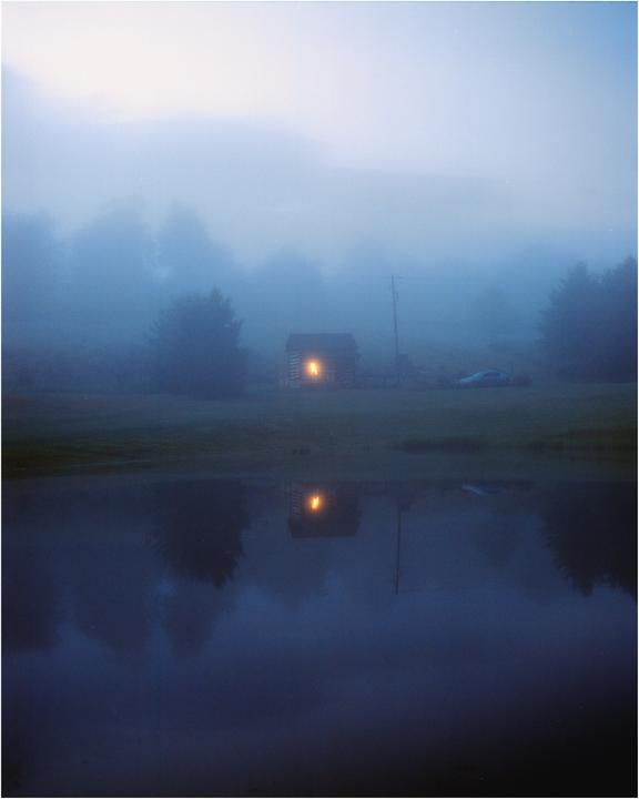 A small cabin with a lit window is reflected in a body of water on a foggy night.