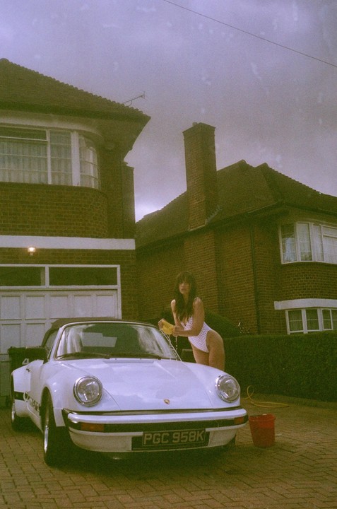 A woman in a white swimsuit washes a white convertible car in front of a brick house.