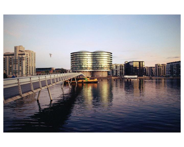 A pedestrian bridge extends over water towards a city skyline with a prominent modern building reflecting the golden hour light.