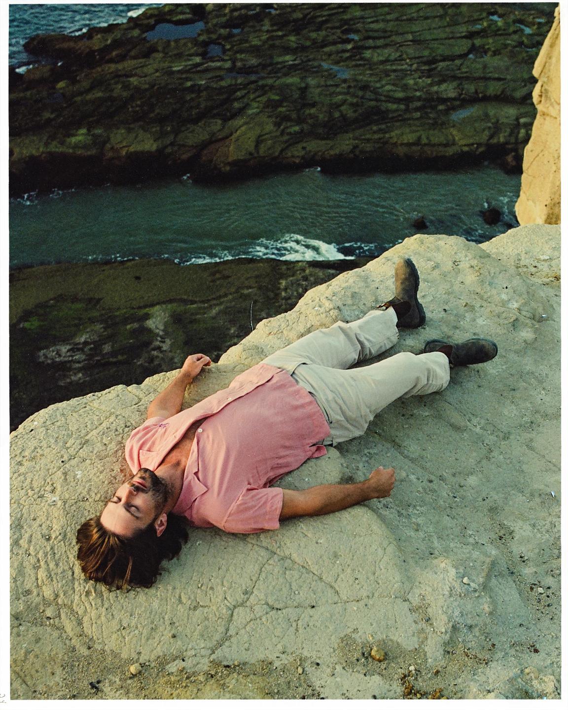 A man in a pink shirt and light pants lies on a rock overlooking the ocean.