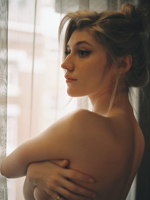A woman with her arms crossed looks out a window covered by a sheer curtain.