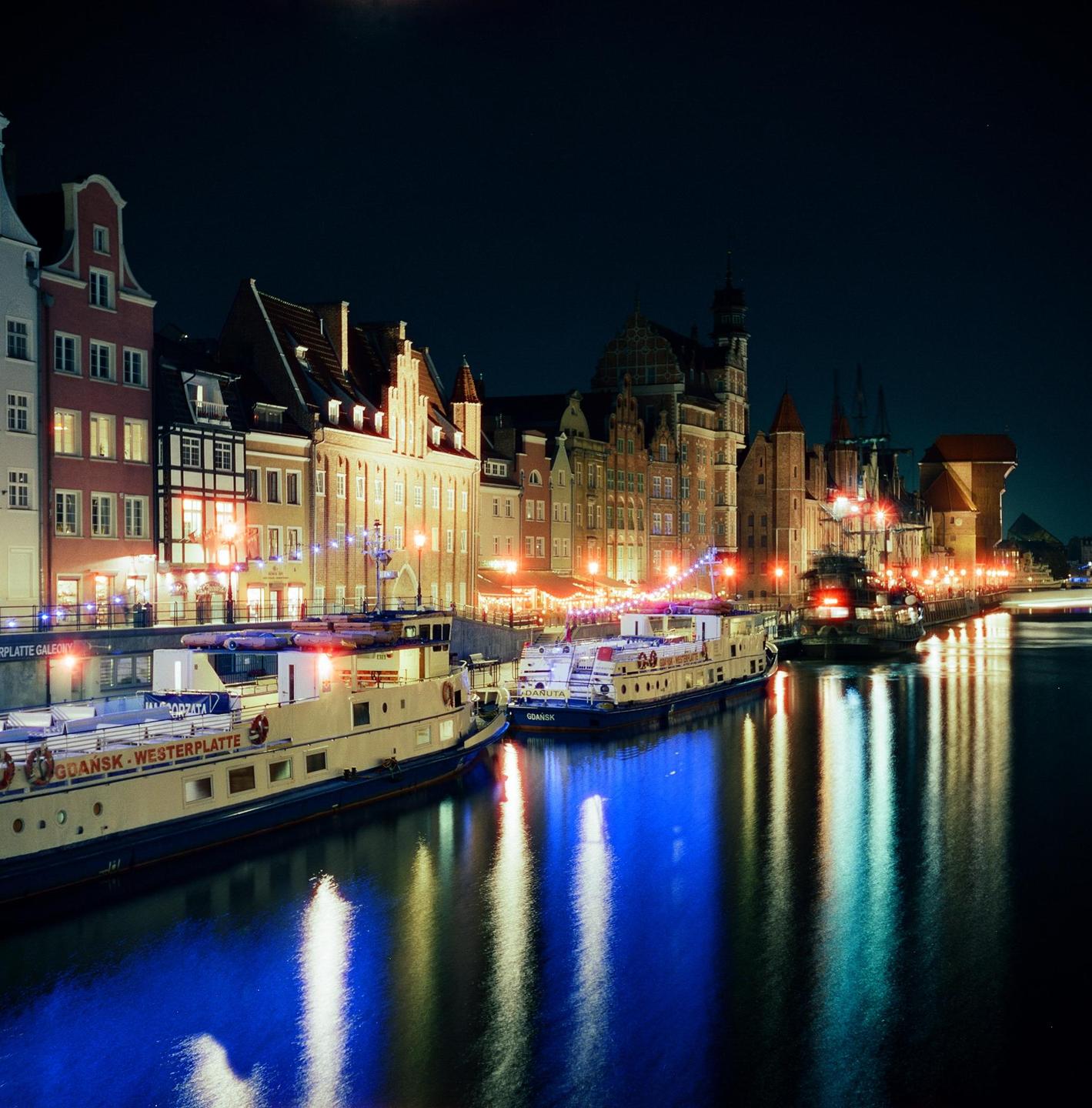 A long exposure captures the illuminated waterfront buildings and boats on the water at night.