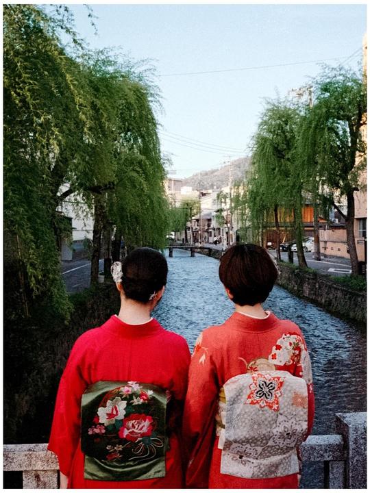 Two women in kimonos stand on a bridge overlooking a canal lined with trees and buildings.