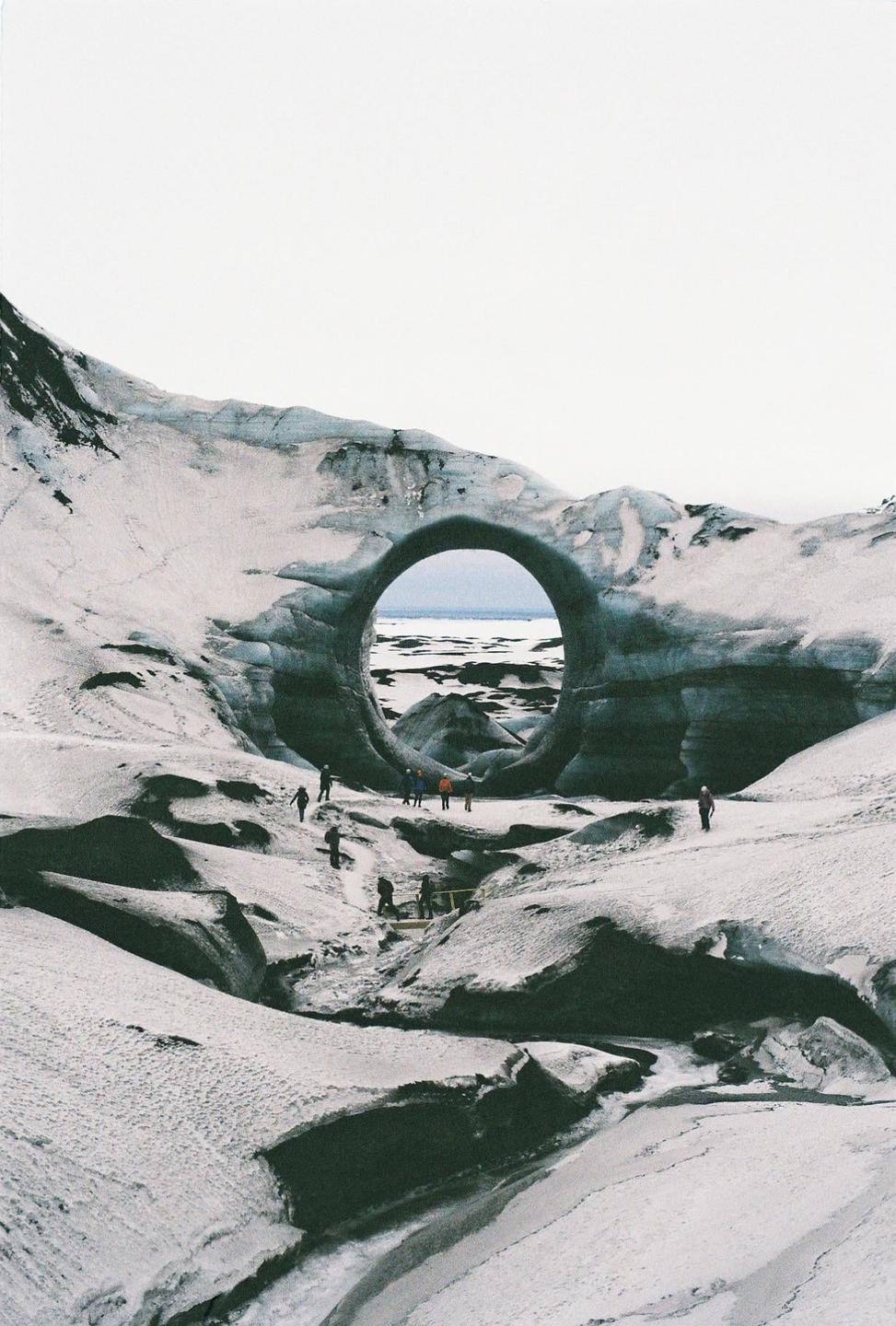 A group of people walk through a snowy landscape with a large circular ice formation in the background.