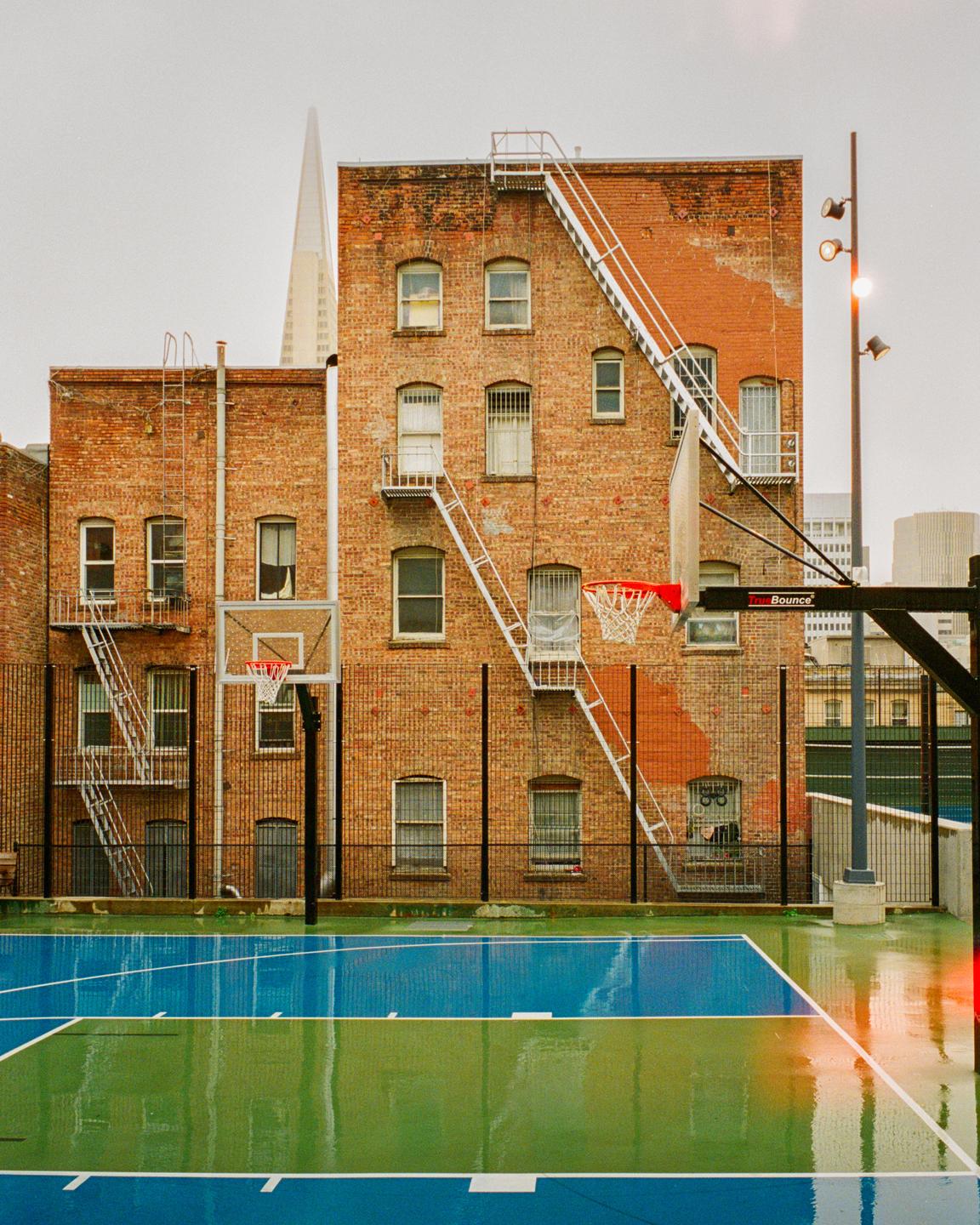 A wet basketball court with blue and green sections is in front of a brick building with fire escapes.
