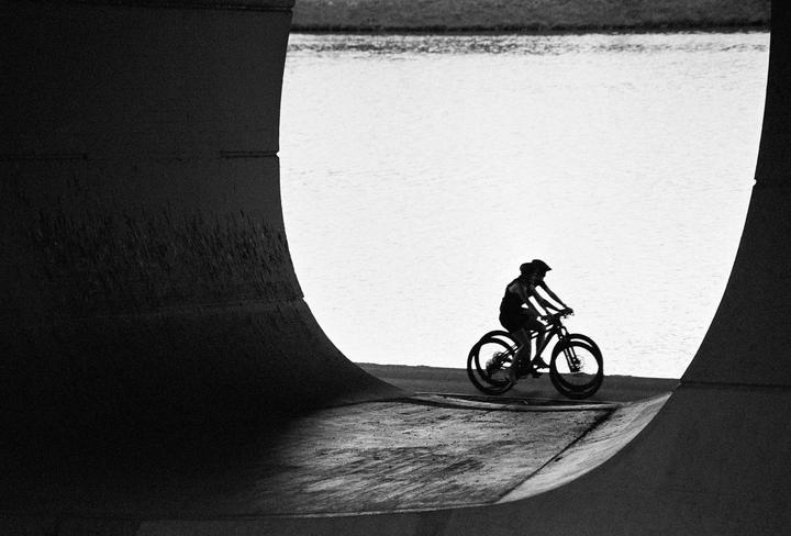 Two cyclists ride bicycles on a concrete path next to a body of water, framed by a dark concrete structure.