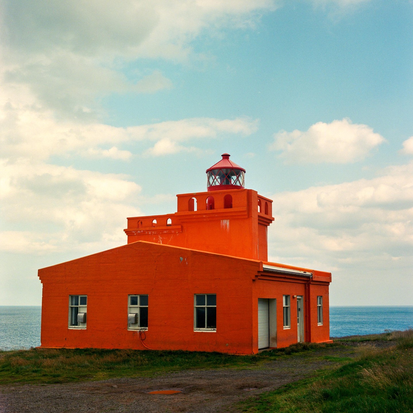 An orange lighthouse stands on a grassy hill overlooking the ocean under a cloudy sky.