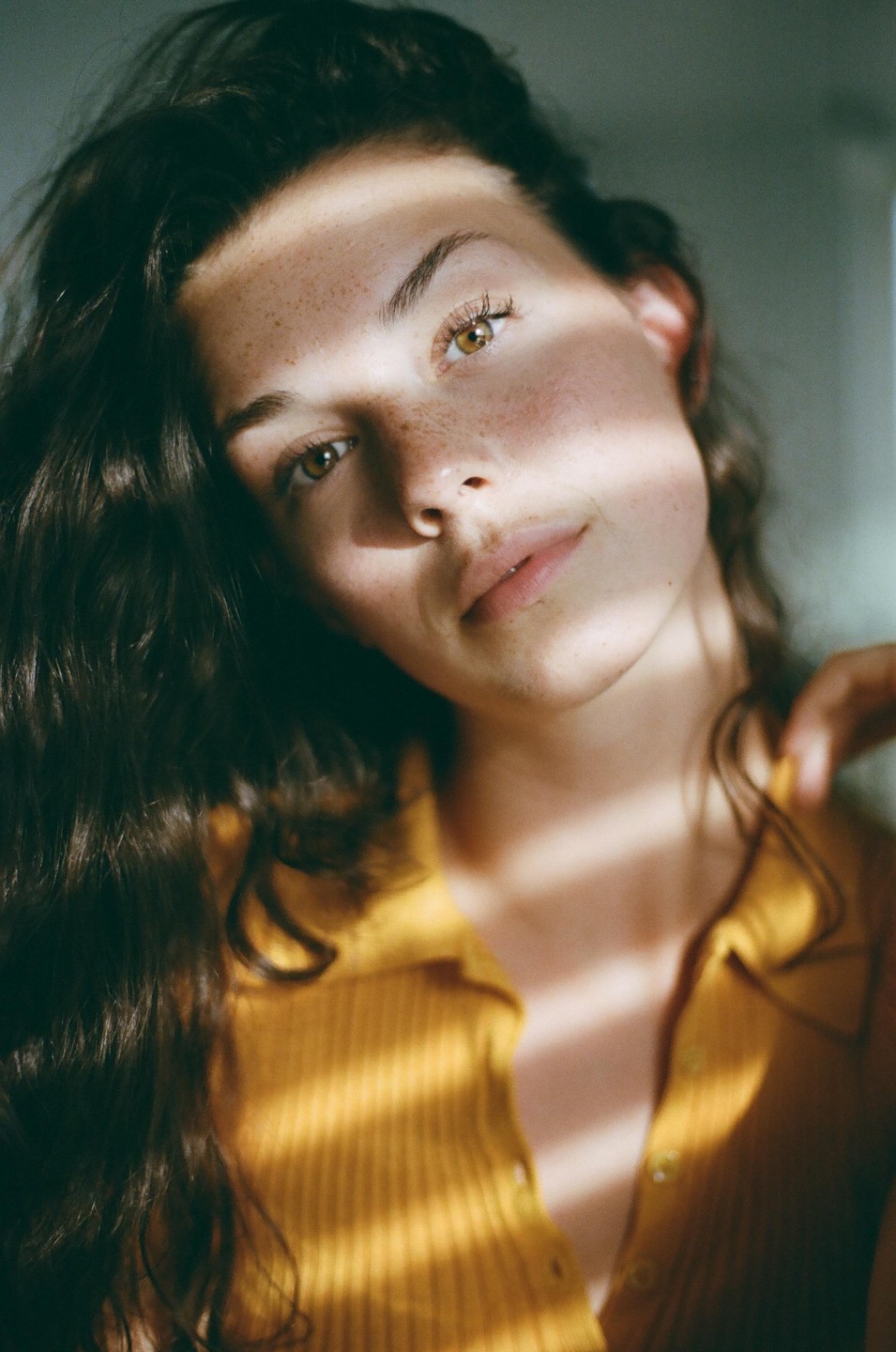 A woman with long dark hair and freckles looks at the viewer, with a shadow across her face.