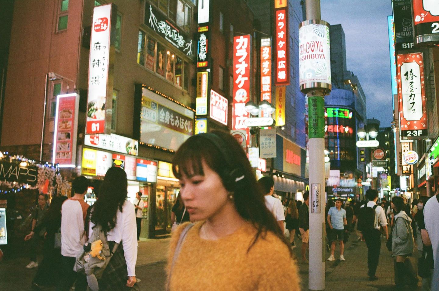 A woman with headphones walks on a busy street lined with neon signs and buildings.