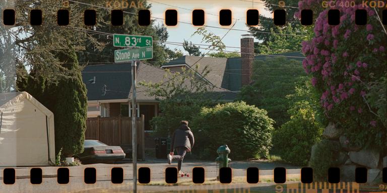 A person on a skateboard moves away from the camera on a residential street with a street sign for N. 83rd St and Stone Ave N.