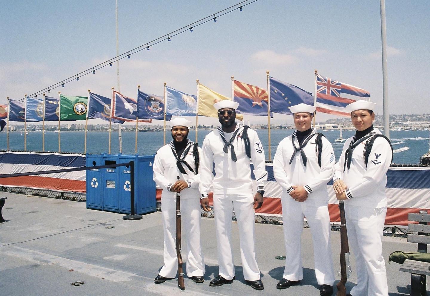 Four sailors in white uniforms stand on a ship's deck with flags and a city skyline in the background.