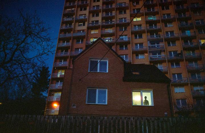 A person stands in a brightly lit window of a small brick house in front of a large apartment building at night.