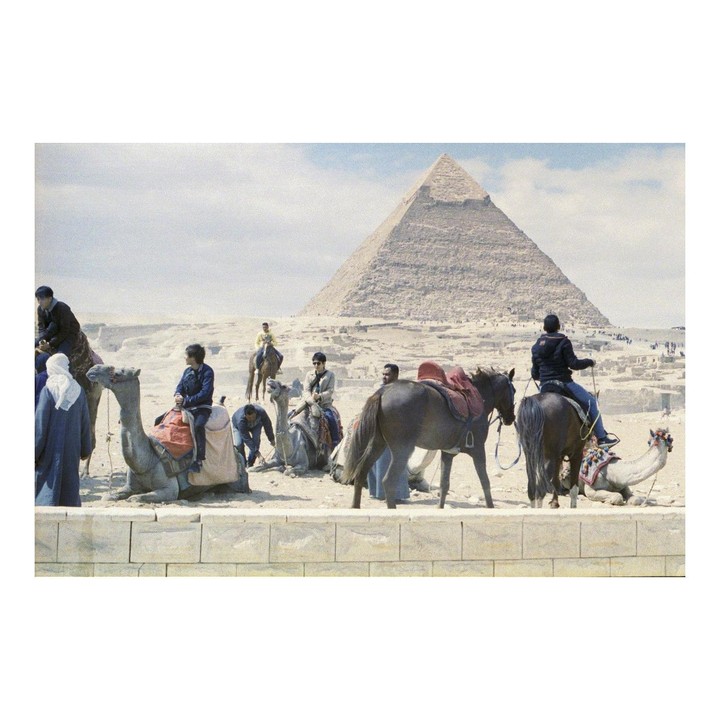 People ride camels and horses in front of a pyramid in a desert landscape.