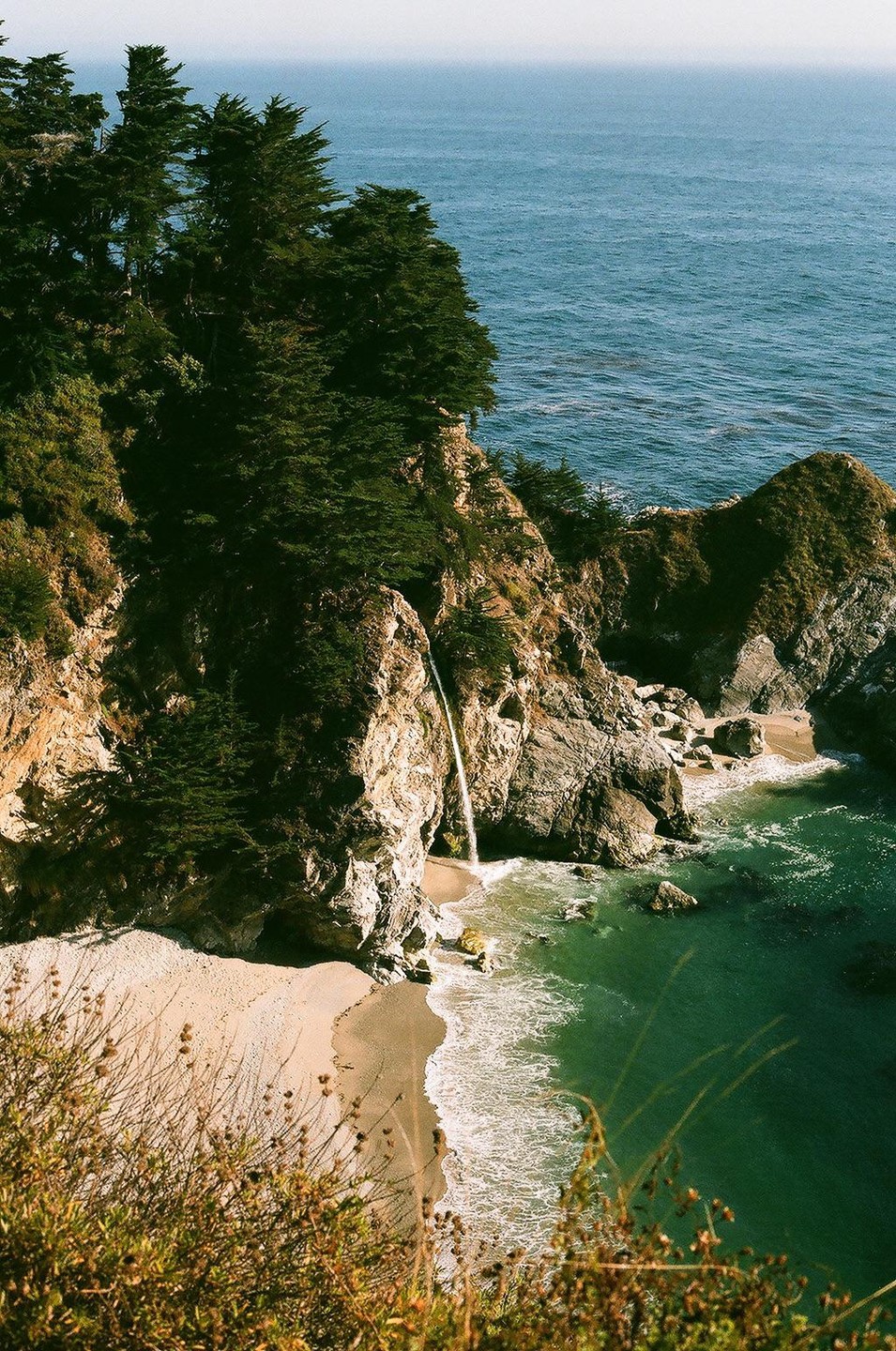 A waterfall cascades onto a sandy beach next to the ocean, surrounded by cliffs and trees.
