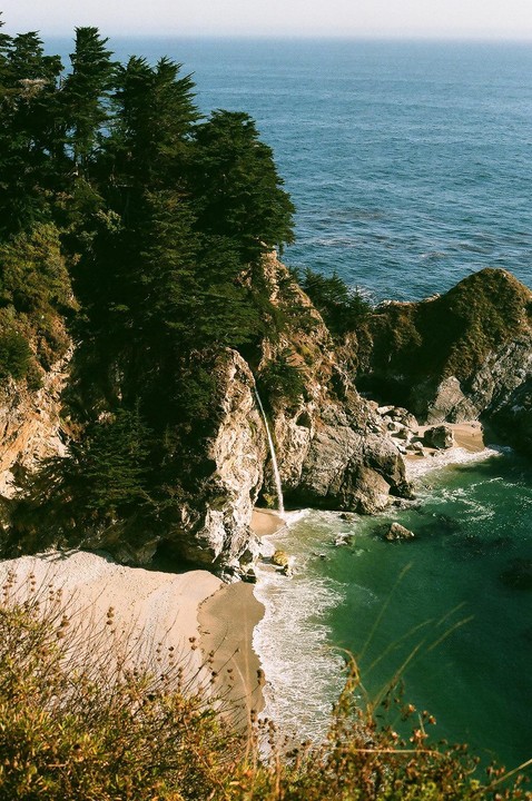 A waterfall cascades onto a sandy beach next to the ocean, surrounded by cliffs and trees.