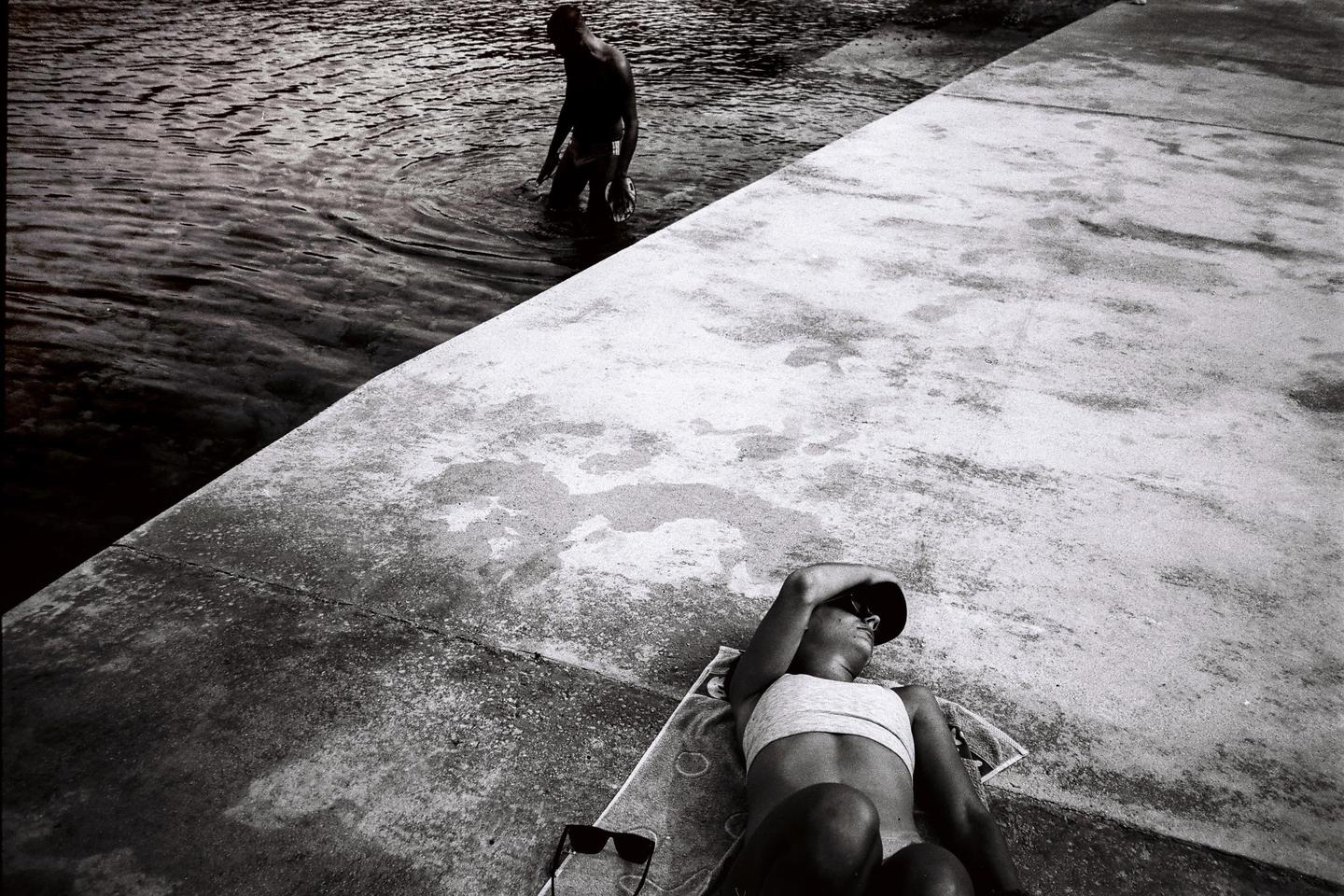 A woman sunbathes on a concrete surface next to water where a man is standing.