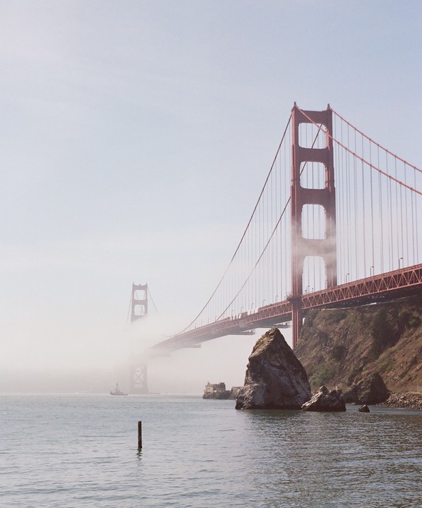 The Golden Gate Bridge is partially obscured by fog over the water, with a rocky shore in the foreground.
