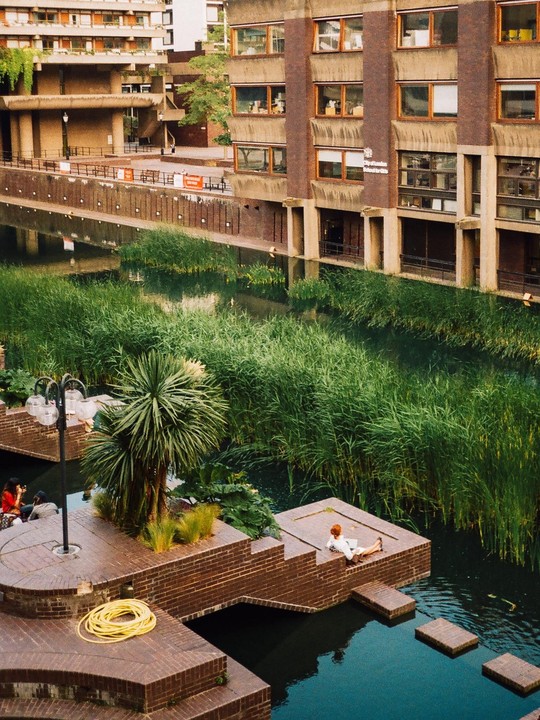 A canal with tall green reeds is surrounded by brick buildings and walkways, with people relaxing on the platforms.
