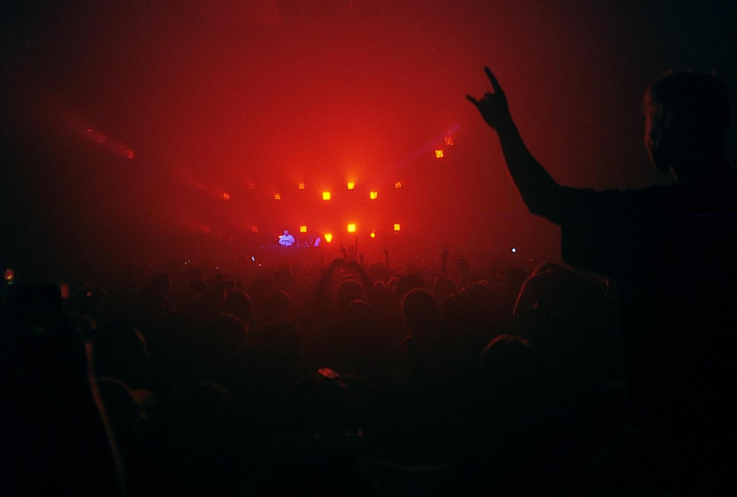 A crowd of people at a concert are illuminated by red stage lights, with one person in the foreground making a hand gesture.