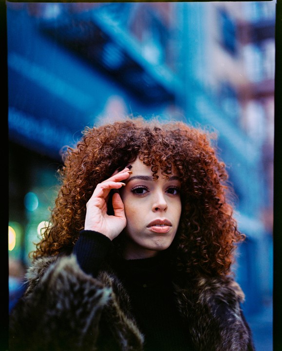 A woman with curly hair and a fur coat looks at the viewer with her hand on her forehead.