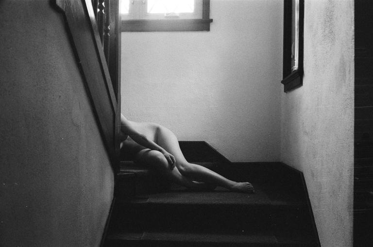 A nude woman is lying on a carpeted staircase next to a window.