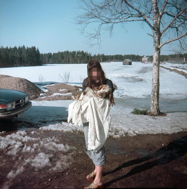 A woman holds a dog wrapped in a white cloth in a snowy landscape with a forest in the background.