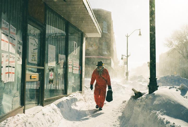 A person in a red jumpsuit and hat walks along a snowy sidewalk next to a storefront with posters.