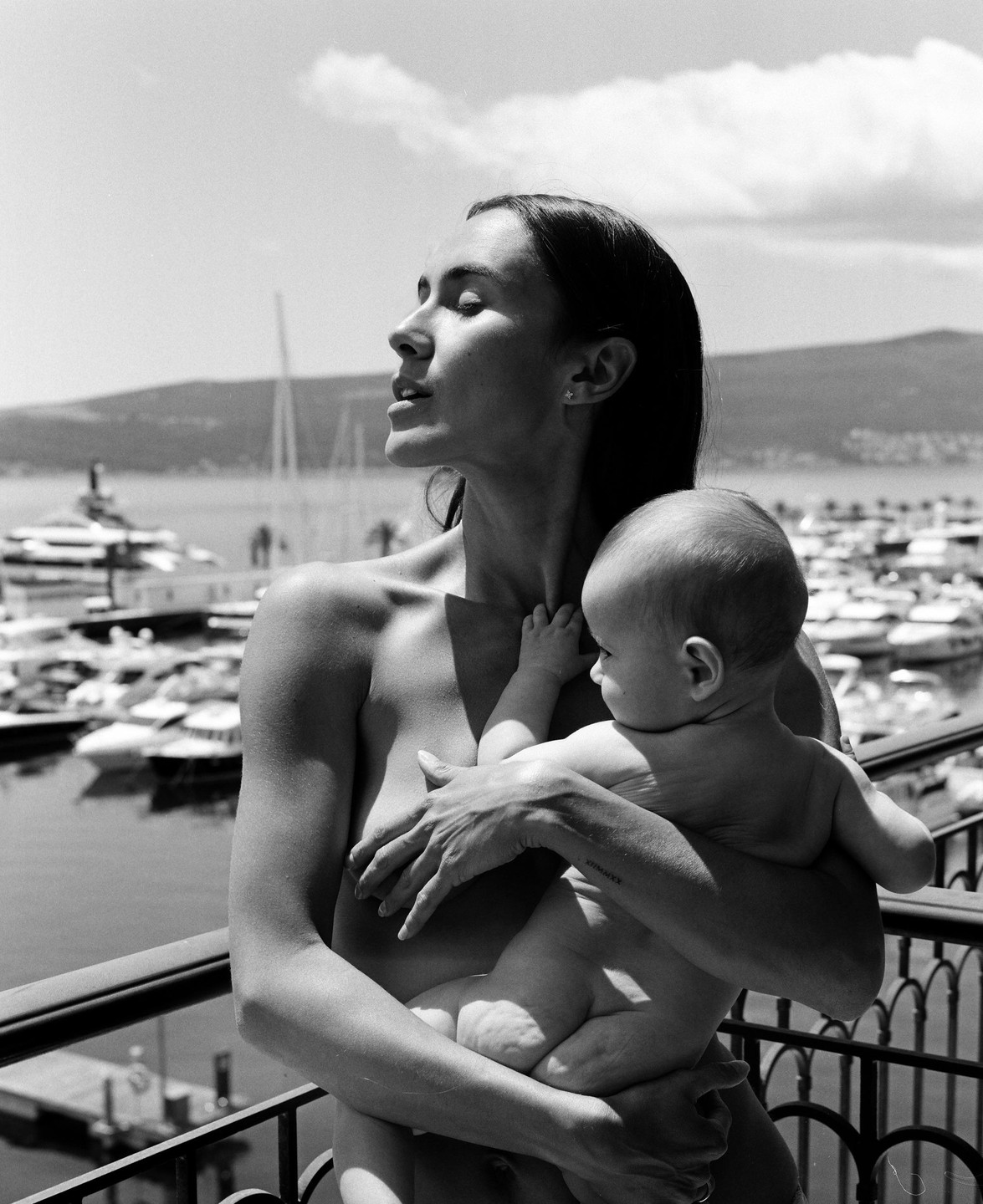 A woman holds a baby on a balcony overlooking a marina with yachts and mountains in the background.