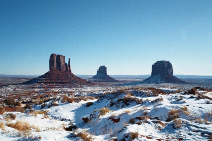 Three large rock formations rise from a snow-covered desert landscape under a clear blue sky.