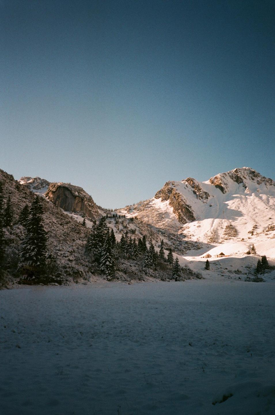 A snow-covered mountain landscape with evergreen trees and small buildings in the distance is visible under a clear sky.