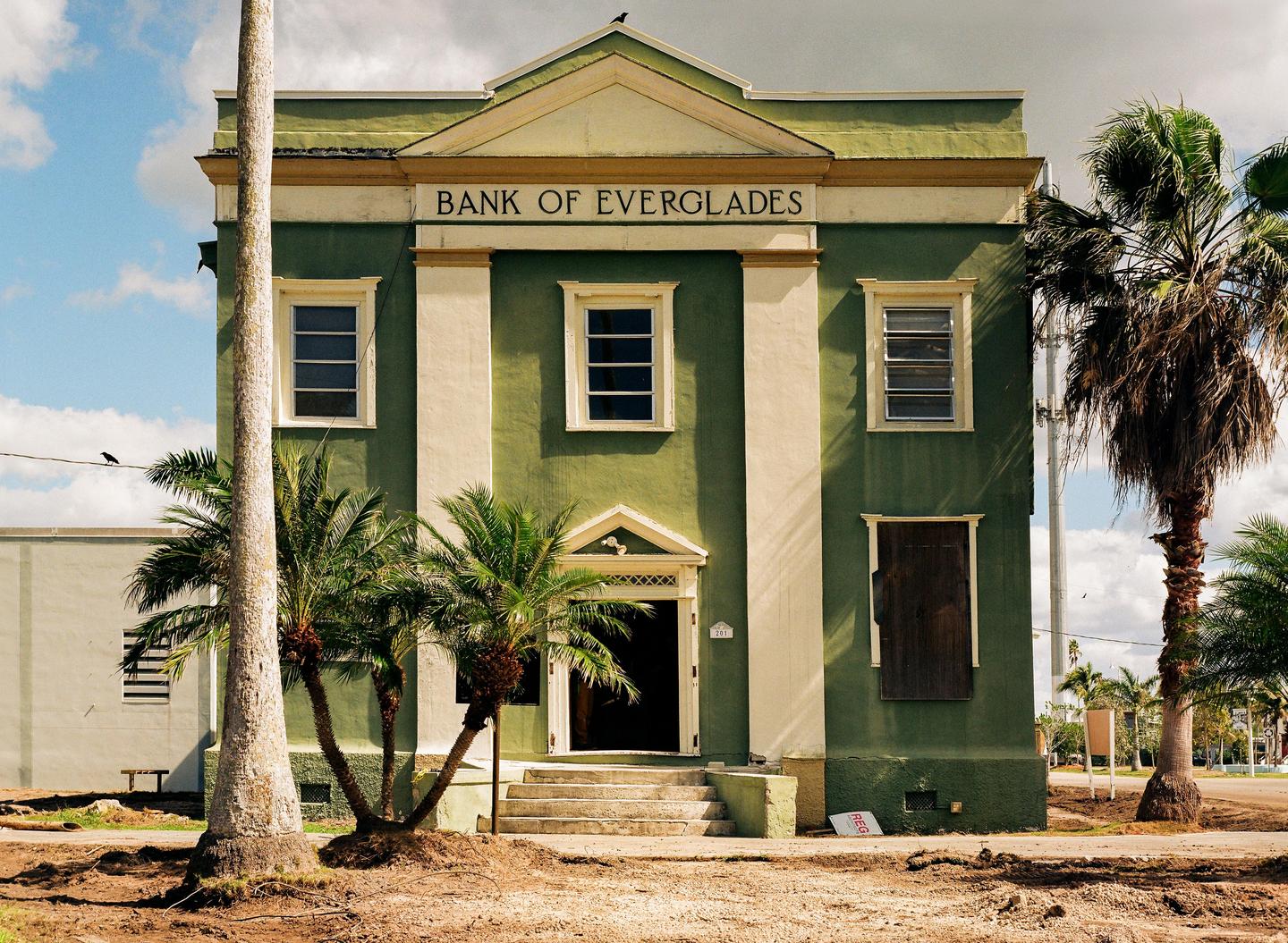 The Bank of Everglades building, painted green and cream, stands under a partly cloudy sky, surrounded by palm trees.