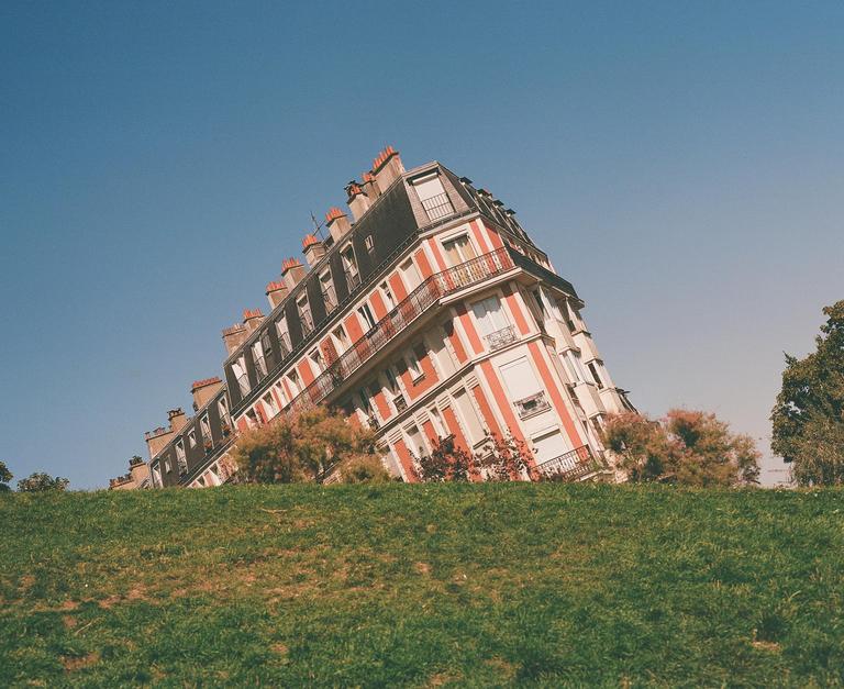 A building with red and white stripes appears to be sinking into a grassy hill under a clear blue sky.