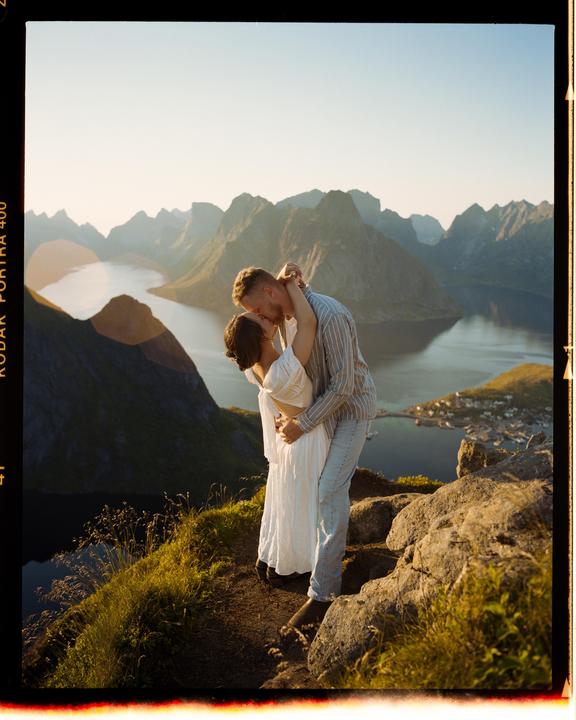 A couple kisses on a mountain overlooking a fjord and a small village.