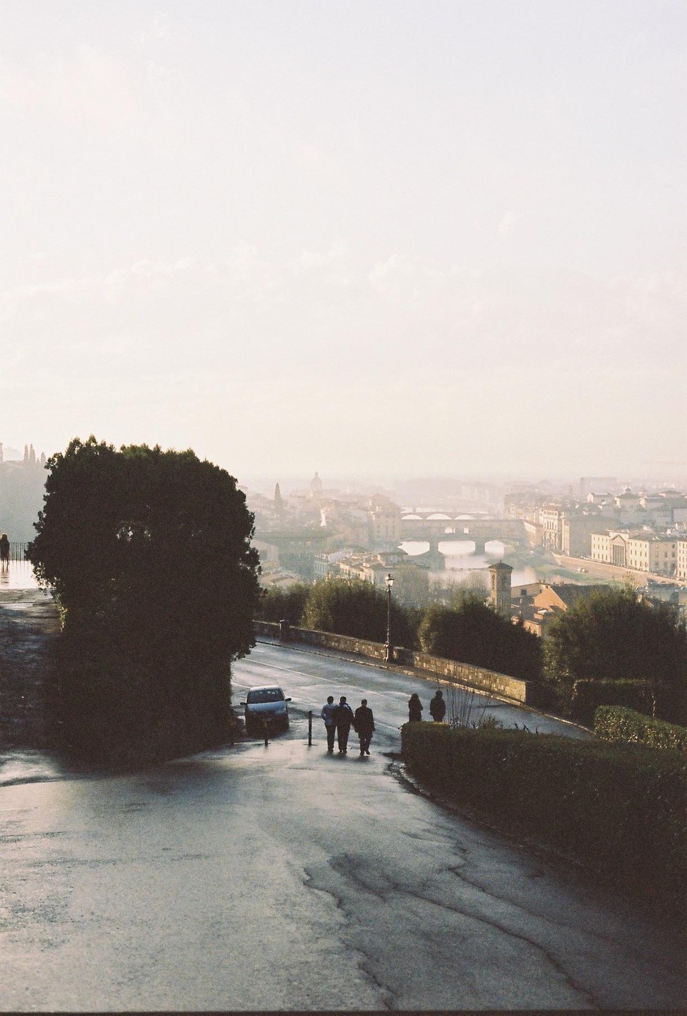 A group of people walk down a wet road overlooking the city of Florence with a bridge over a river in the distance.