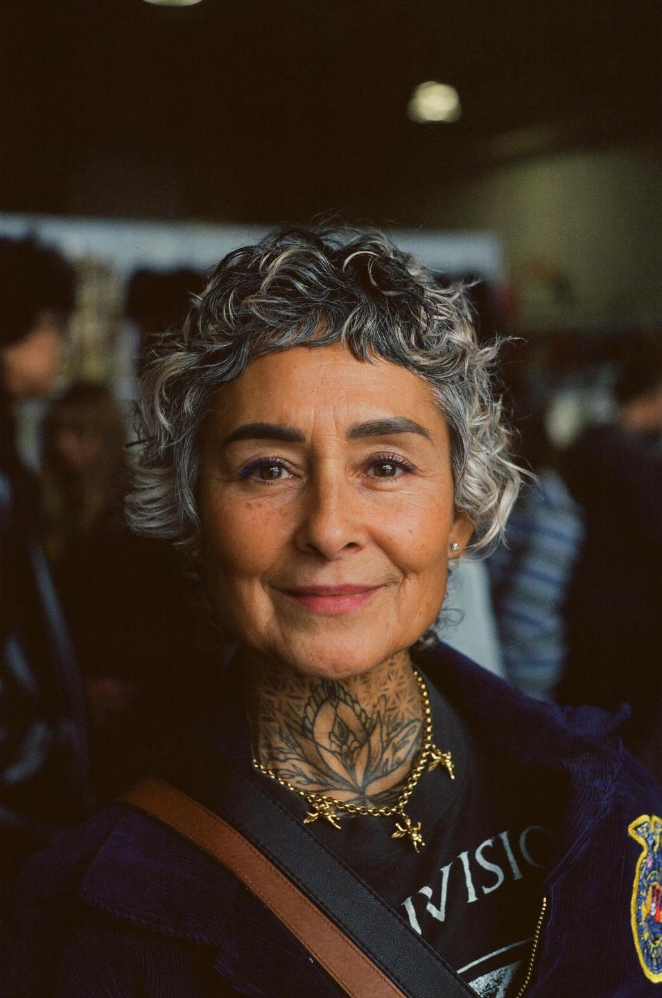 A woman with short curly hair and a tattooed neck smiles at the viewer.