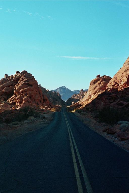 A road with double yellow lines leads through a desert landscape with red rock formations and mountains under a clear blue sky.