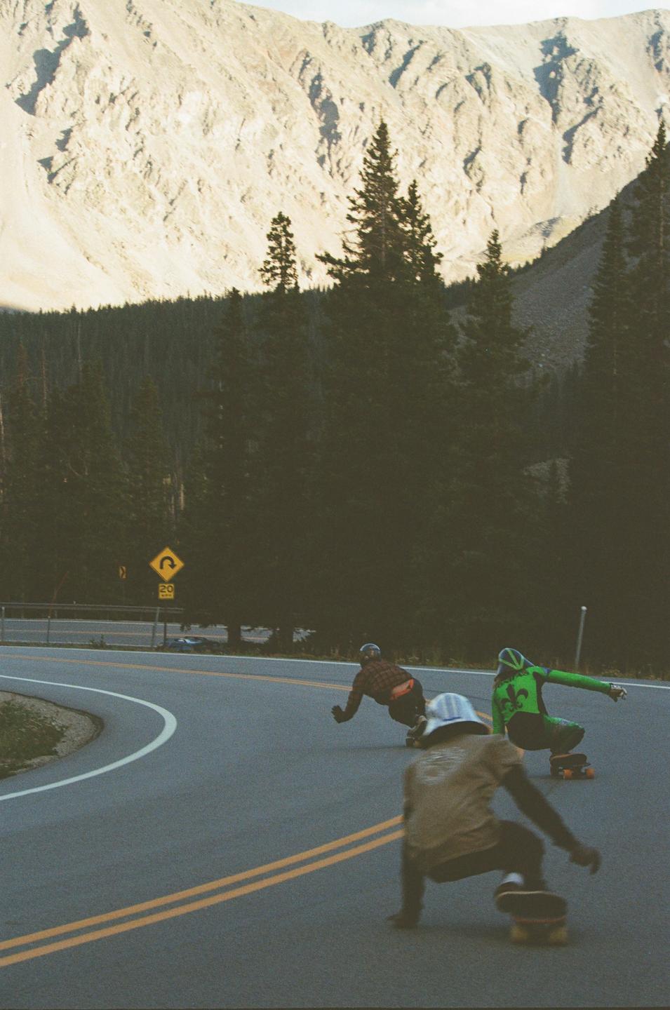 Three men on skateboards race down a winding mountain road lined with pine trees and a rocky mountain.