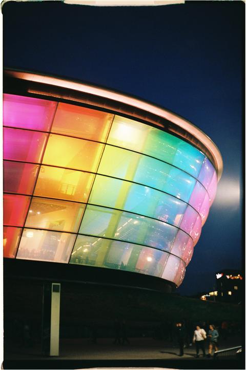 A building with a curved, rainbow-colored glass facade stands against a dark blue night sky.