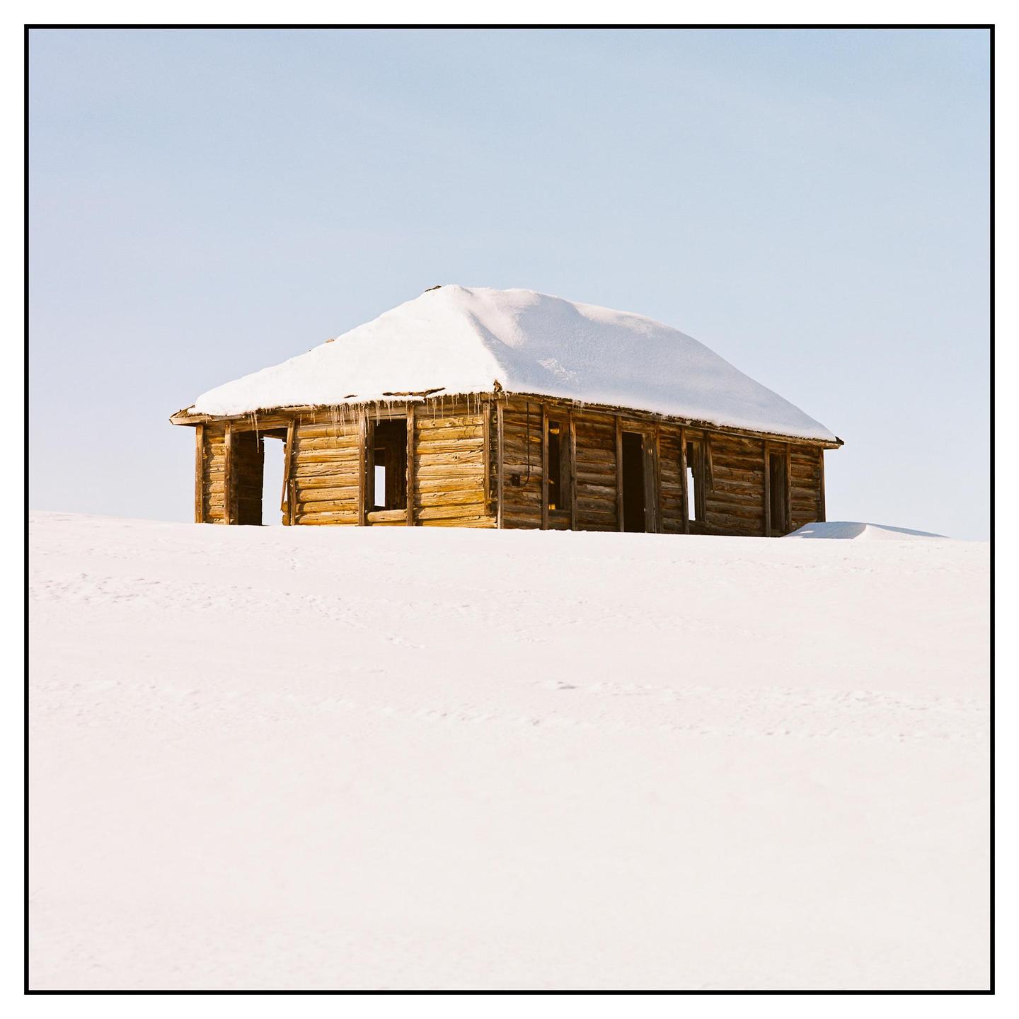 A wooden cabin with a snow-covered roof stands in a field of snow under a clear sky.