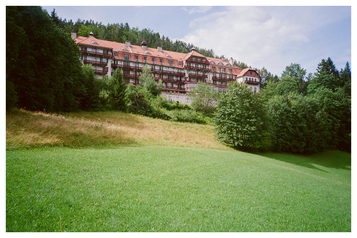 A large, multi-story building with a red roof and many balconies is nestled into a hillside covered in trees and grass.