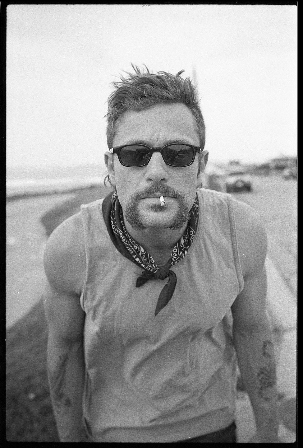 A man with sunglasses, a bandana, and a cigarette in his mouth looks directly at the camera.